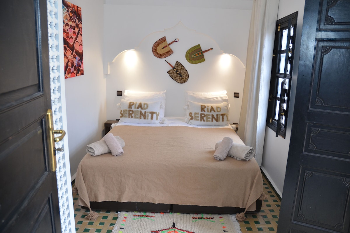 Un llit ben fet amb un estovat beix i dos coixins amb la inscripció 'Riad Serenity', una catifa amb dibuixos i una porta amb manilla de bronze.