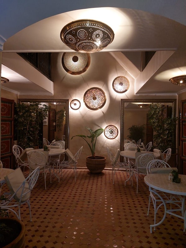 An outdoor dining area with white metal chairs and tables, a central potted plant, and decorative wall sconces.
