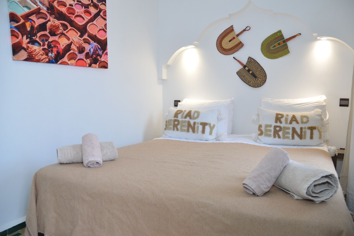 Een netjes opgemaakt bed met twee kussens met de tekst 'PIAD SERENITY' en een aan de muur hangend kunstwerk dat een kleurrijke scène voorstelt.
