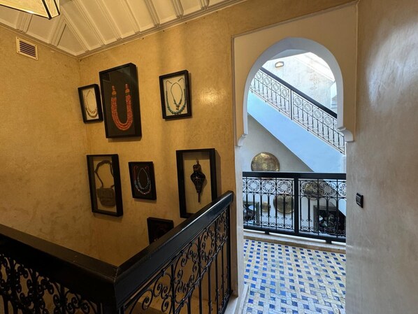 A hallway with framed artwork on the walls, a black wrought iron railing, and a blue and white tiled floor leading to a staircase. - Riad Dar Serenity (Marrakech)