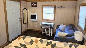 A cozy room with a wooden ceiling, a blue sofa with cushions, a small table with a lamp, a window, and a door. - Cozy Warm Creekside Escape w/ WiFi & Hot Shower (Purlear)
