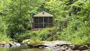 A rustic cabin with a screened porch, surrounded by lush greenery and a flowing stream. - Cozy Warm Creekside Escape w/ WiFi & Hot Shower (Purlear)
