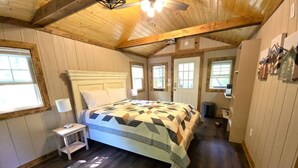 A bedroom with a wooden ceiling, a bed with a patterned bedspread, a bedside table with a lamp, and a window. - Cozy Warm Creekside Escape w/ WiFi & Hot Shower (Purlear)