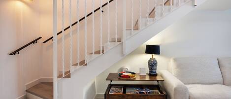 Una escalera con barandillas blancas y un pasamanos de madera, un sofá beige, una mesa de centro de madera con objetos decorativos, y un aplique de pared.