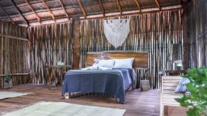 A rustic wooden room with a bed, a woven macrame wall hanging, and a small table with a lamp. - Jungla & Estrellas (Coba)