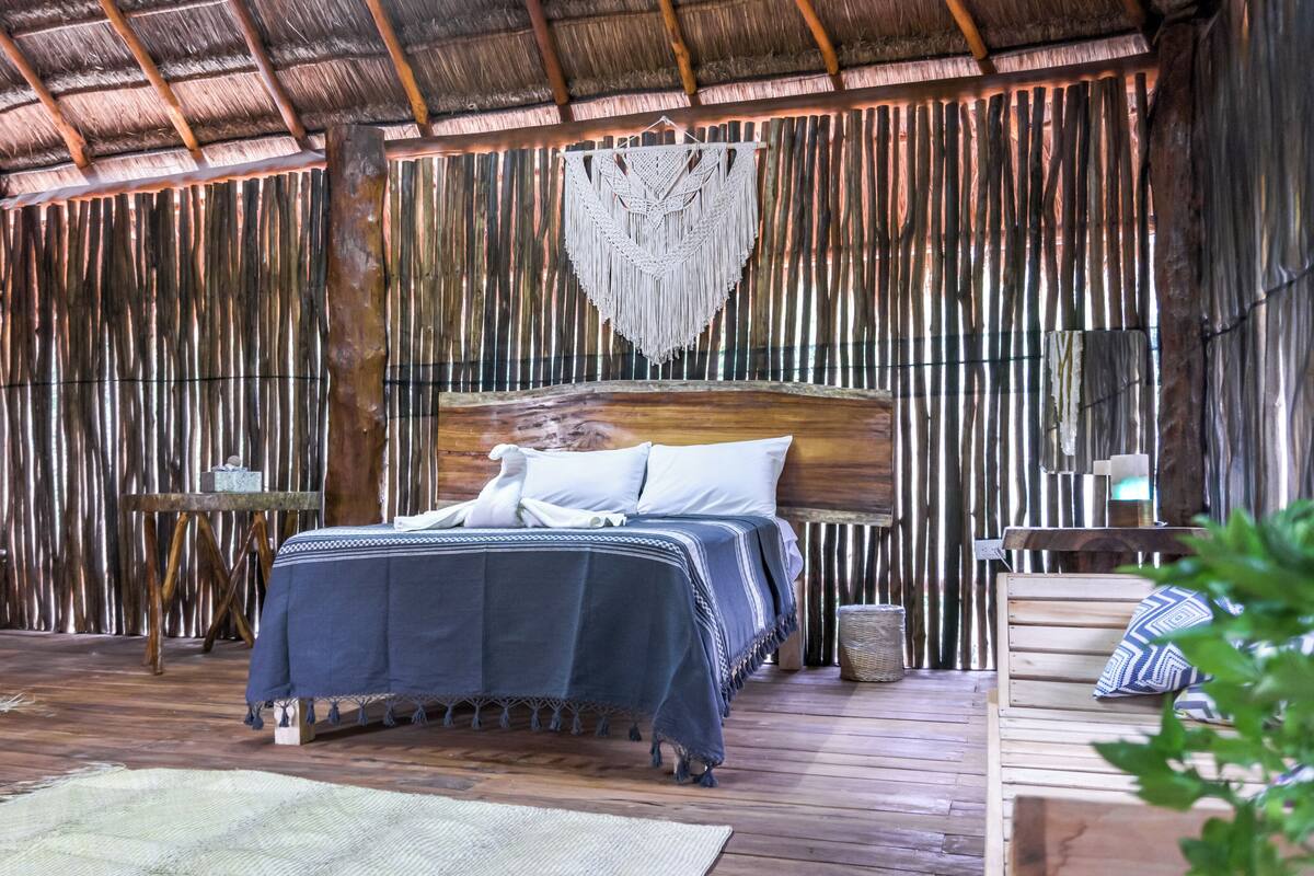 A rustic wooden room with a bed, a woven macrame wall hanging, and a small table with a lamp.