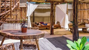 A wooden deck with a round table, wicker chairs, and a potted plant. There are wooden structures and a thatched roof in the background. - Jungla & Estrellas (Coba)