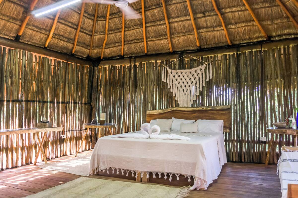 A room with a bed, wooden floors, a ceiling fan, and a thatched roof.