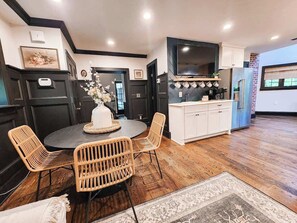 A modern kitchen with dark cabinetry, a large TV, and a dining area with wicker chairs and a round table.