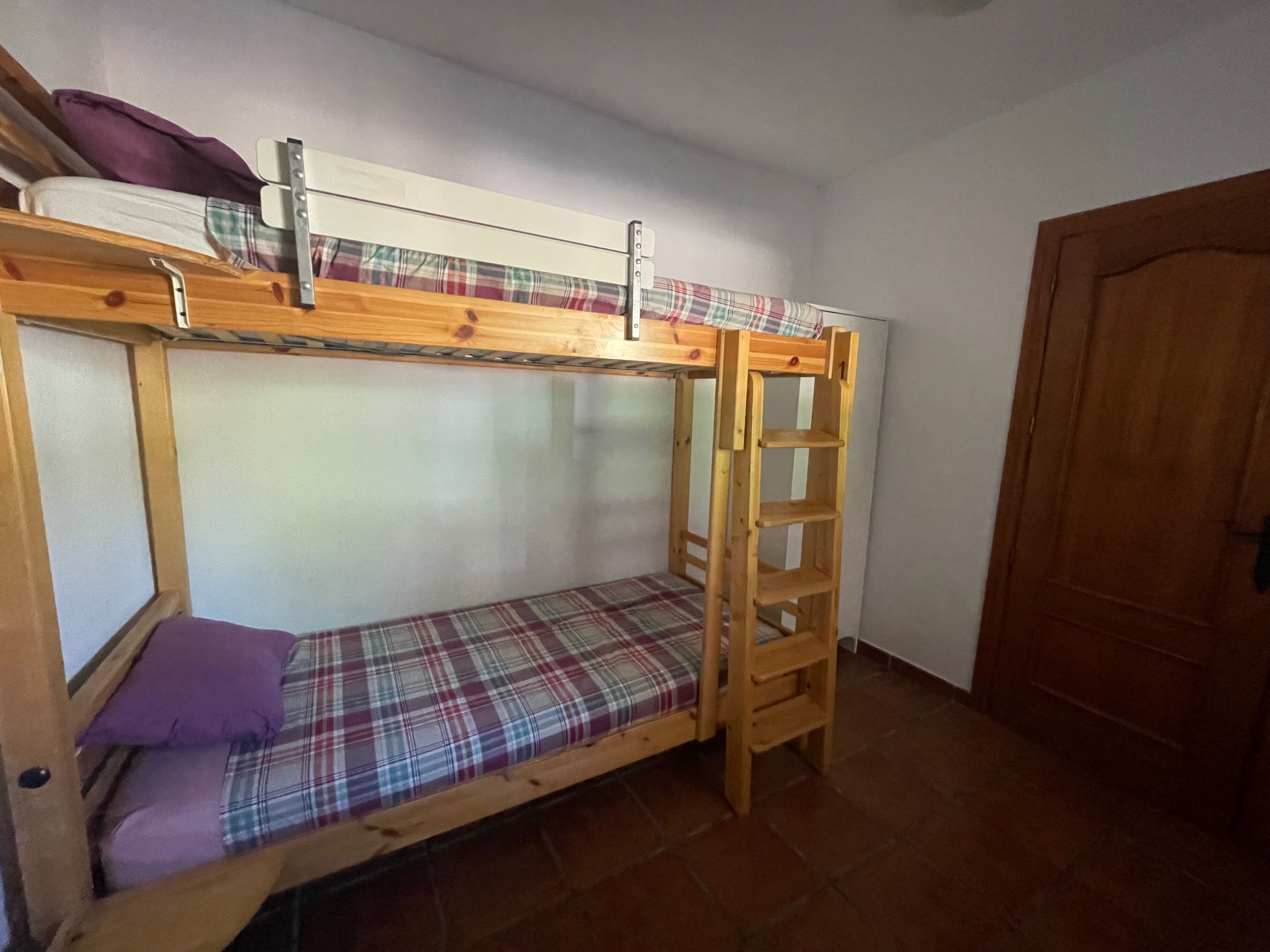 Een stapelbed met een houten trapje, een geruit matras en een paars kussen.