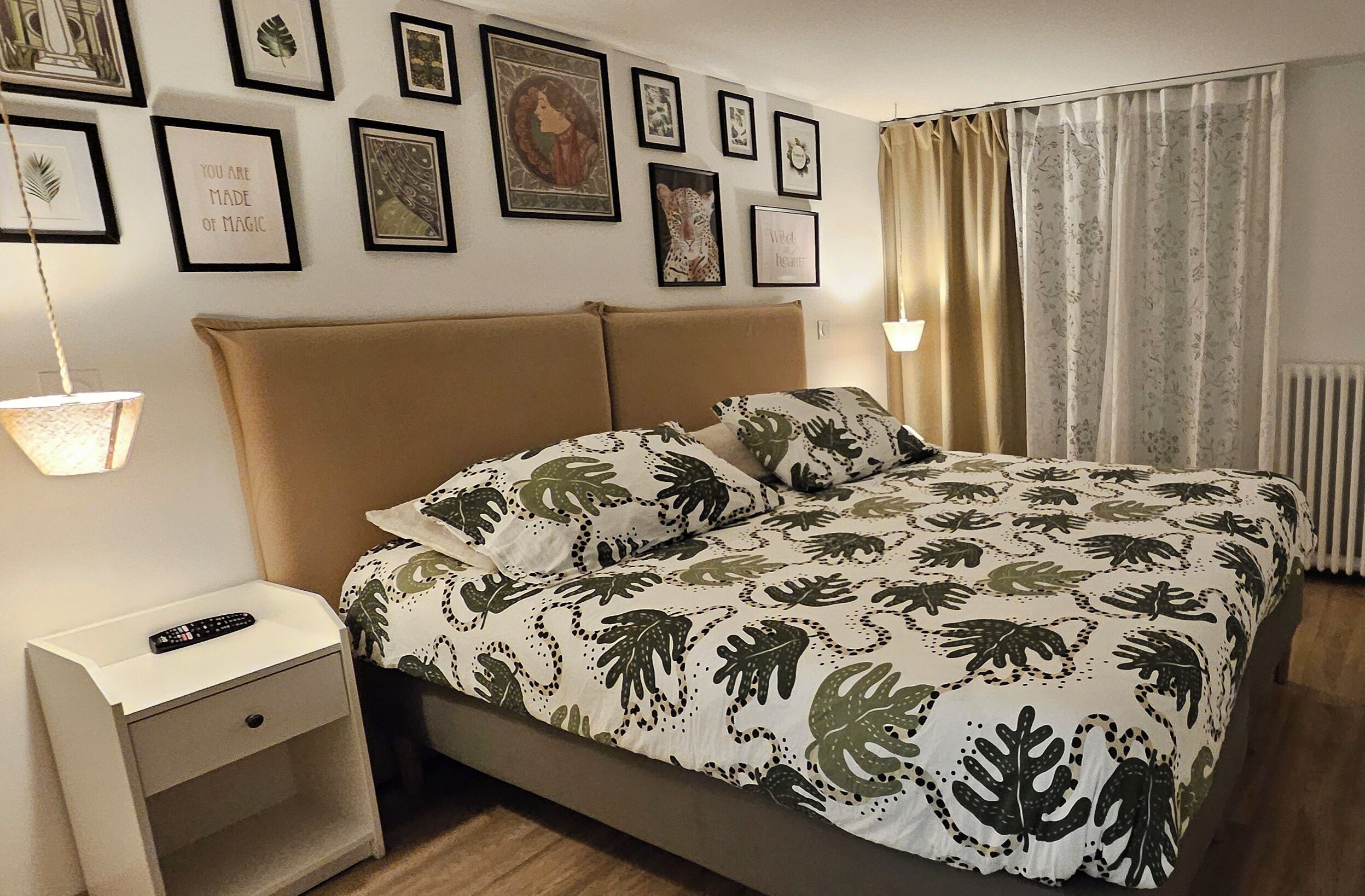A bedroom with a bed, a nightstand, a wall with framed pictures, and a window with curtains.