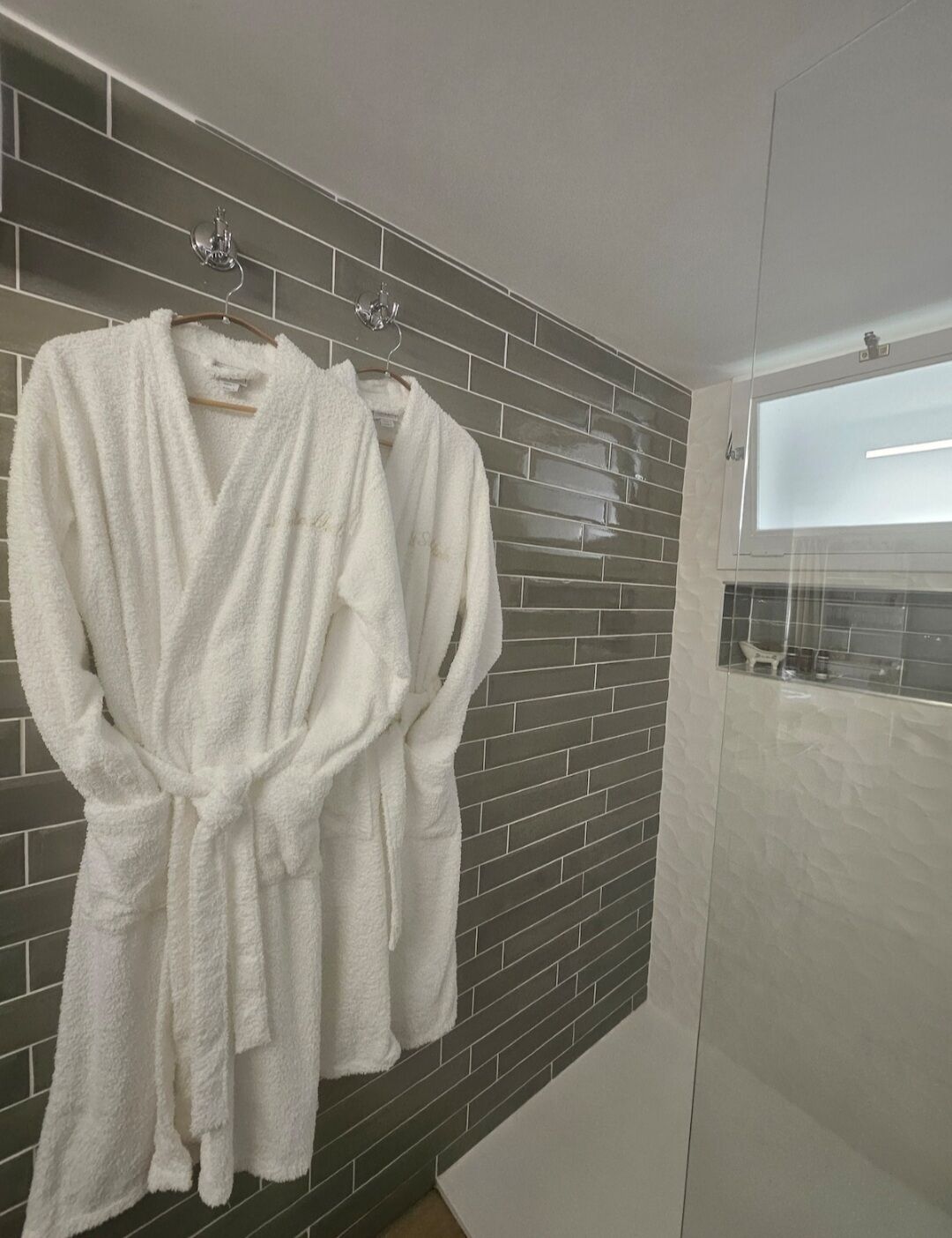 Een badkamer met een grijze betegelde muur, een douchecabine met glazen wanden, en twee witte badjassen die aan een rek hangen.