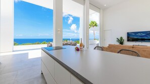 Een moderne woonkamer met een groot raam met uitzicht op de oceaan, een eethoek met terrasmeubels en een flatscreen aan de wand.