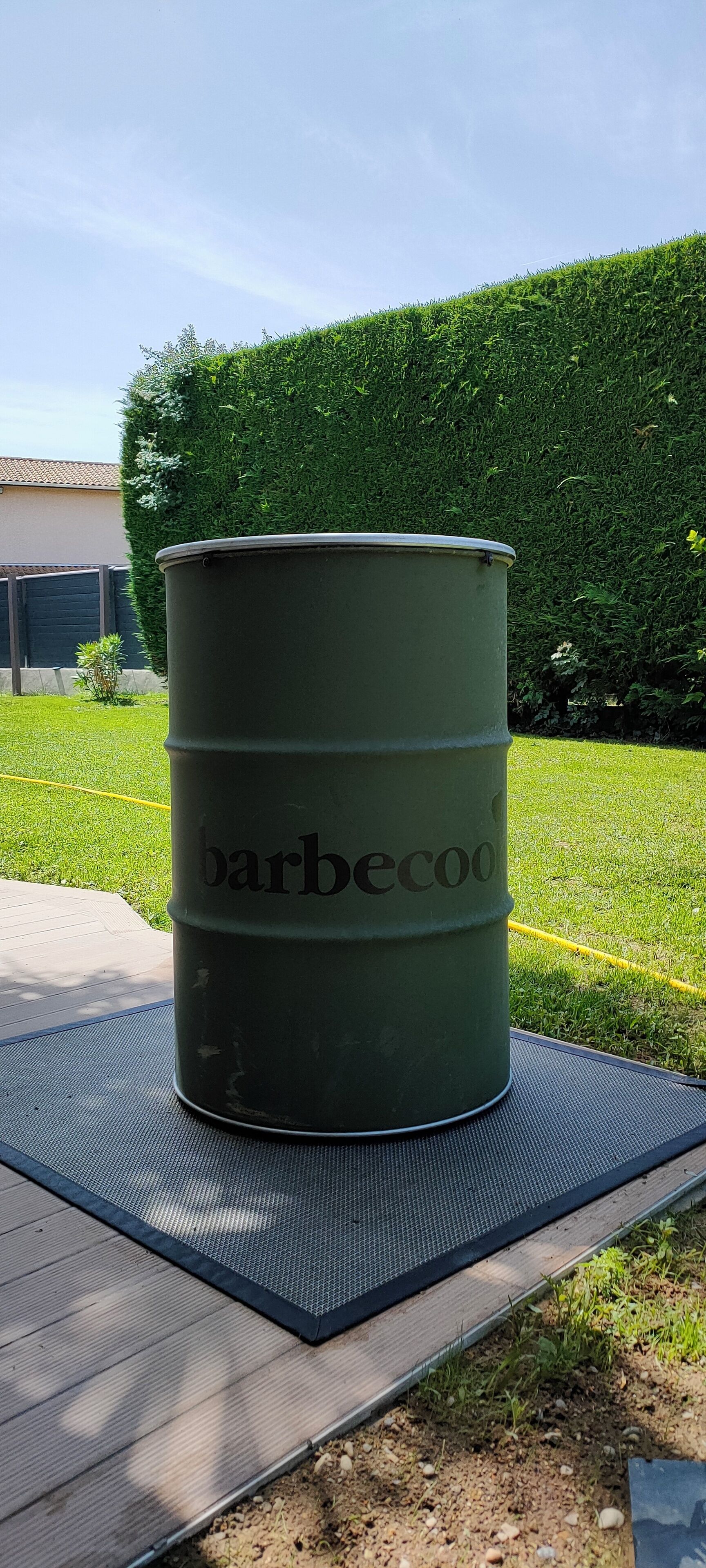 Un barril verd amb el text "barbecoo" a sobre, col·locat sobre un estora a l'exterior.