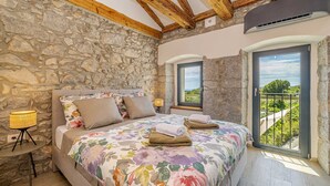 A bedroom with a stone wall, wooden beams, a bed with floral bedding, two windows, and a balcony with a view. - Holiday house RURAL HOME FRANE (Kornić)