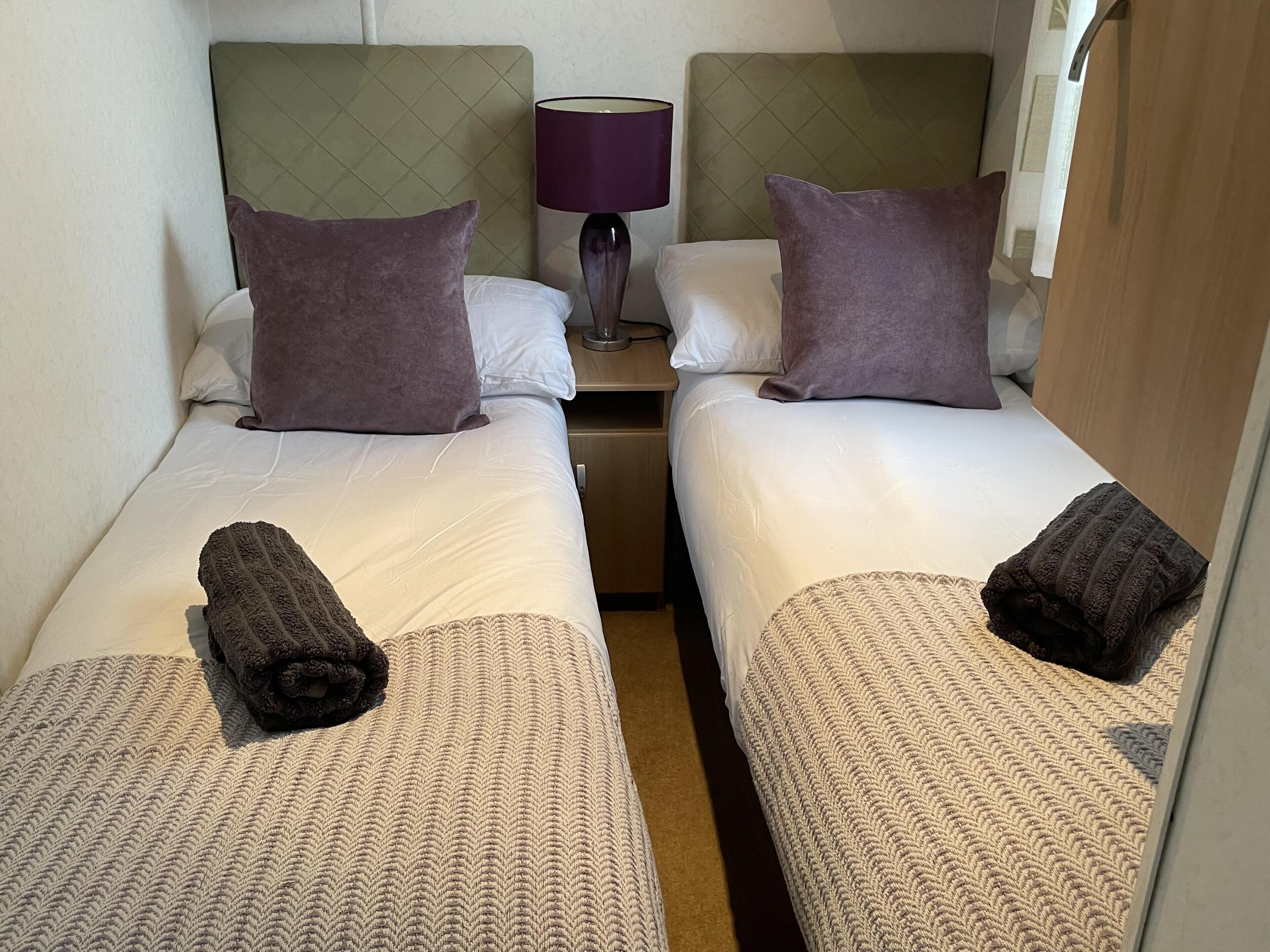 Ein Hotelzimmer mit zwei Einzelbetten, jeweils mit einem Kopfkissen und einem Handtuch ausgestattet. Zwischen den Betten befindet sich ein Nachttisch mit einer Lampe.
