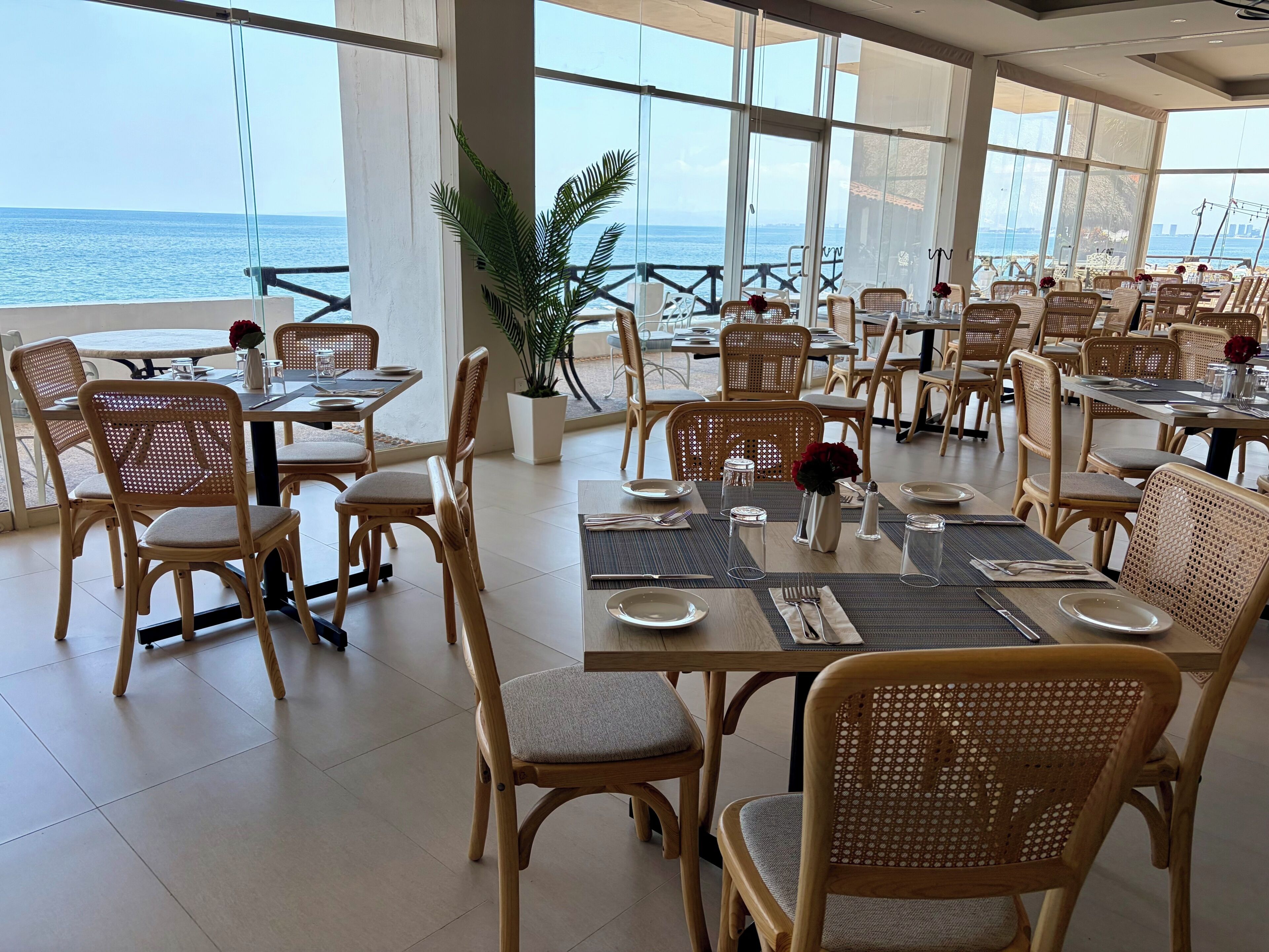 Un restaurant avec des chaises et des tables en osier, de grandes fenêtres donnant sur la mer et des plantes en pot.