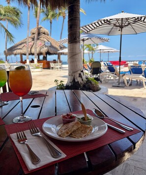 A table with a plate of food, drinks, and cutlery, set against a beach backdrop with umbrellas and palm trees. - Costa Sur All Inclusive Classico Collection by Sonesta (Puerto Vallarta)