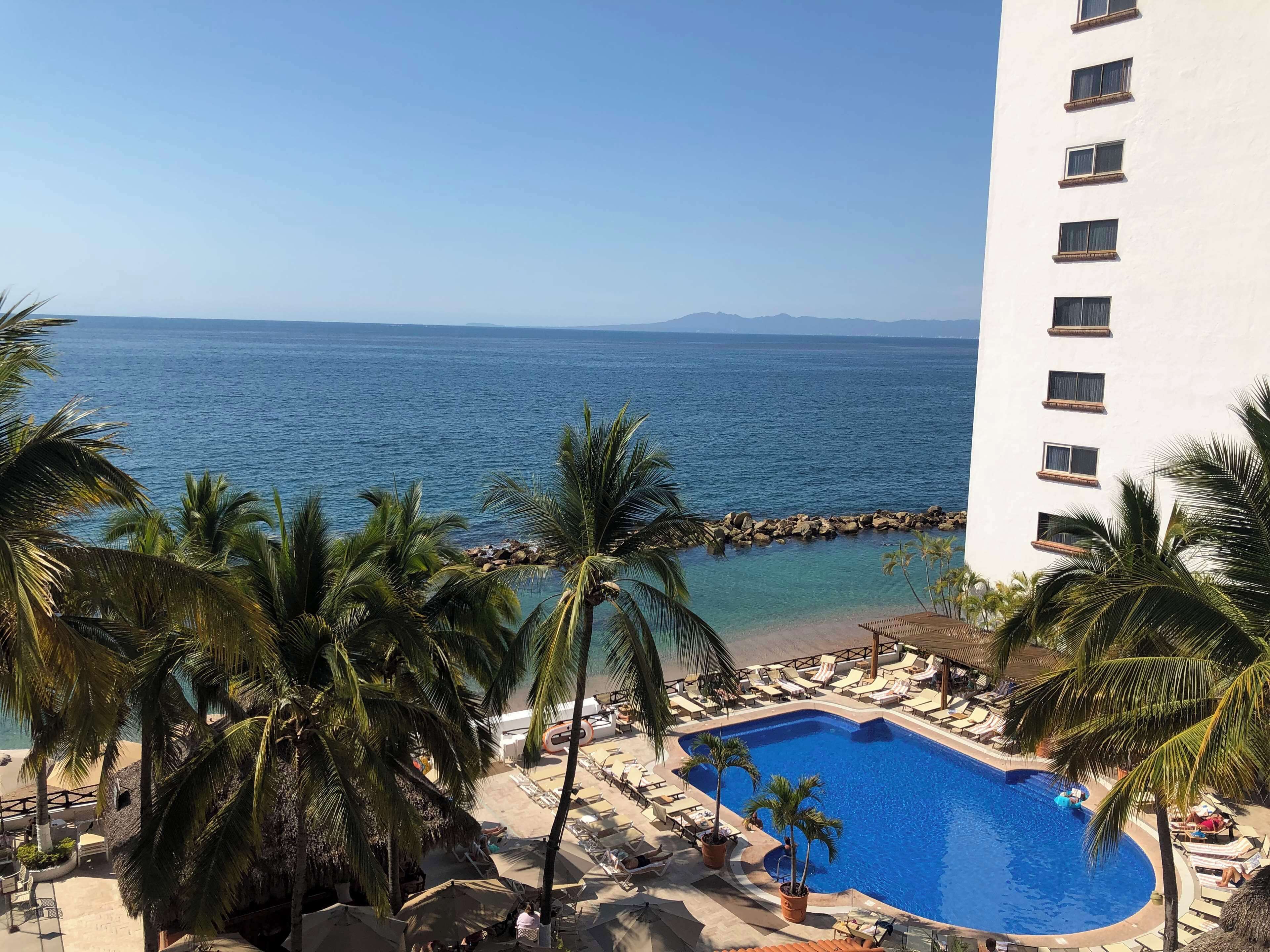 Zwembad van een resort met helderblauw water, omgeven door palmbomen en ligbedden, met uitzicht op een strand en de oceaan.