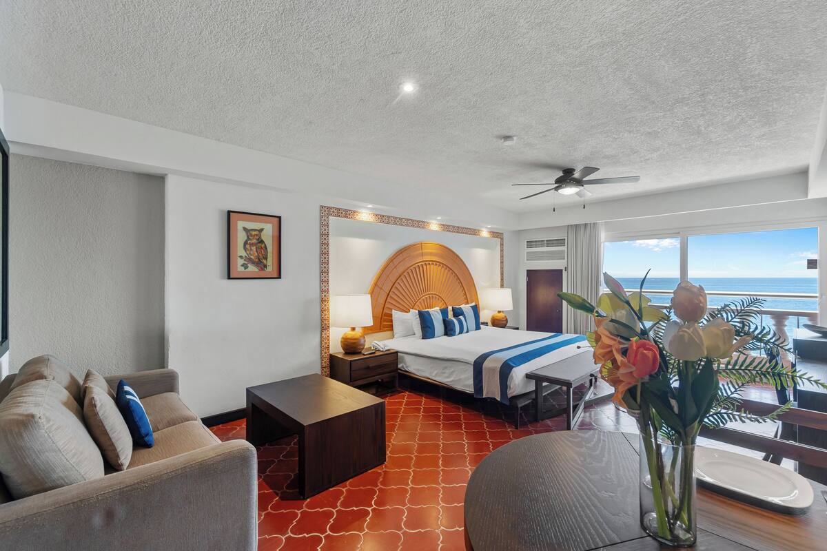 Quarto de hotel com cama, sofá, mesa de jantar e vista para o oceano.