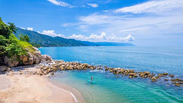A beach with clear turquoise water, a rocky breakwater, and a person paddleboarding. - Costa Sur All Inclusive Classico Collection by Sonesta (Puerto Vallarta)