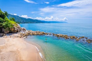 Ein Strand mit klarem türkisblauem Wasser, einem steinernen Wellenbrecher und einer Person, die auf einem Stand-Up-Paddleboard fährt.
