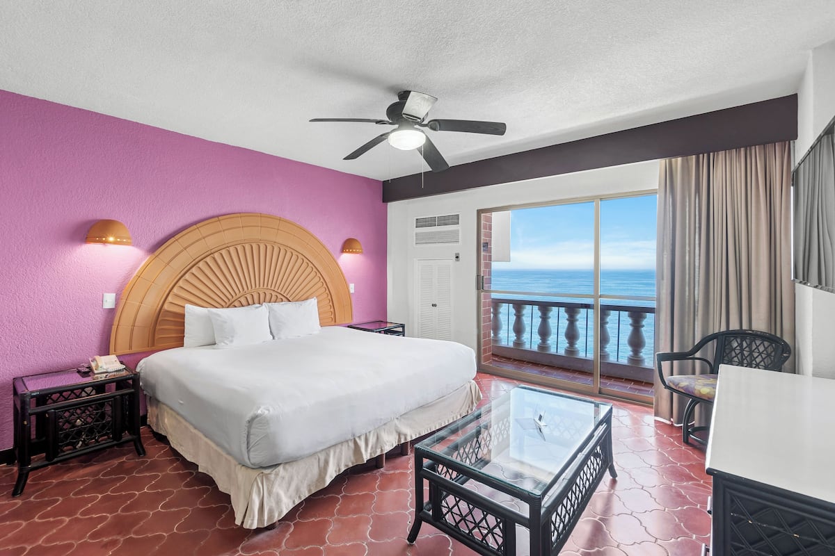 Quarto de hotel com uma cama, um ventilador de teto, uma mesa de vidro, uma cadeira e uma varanda vista para o mar.