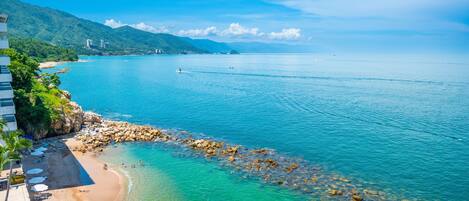 A coastal view with clear turquoise waters, a sandy beach, and rocky outcrops.