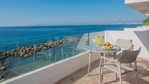 A balcony with a glass railing overlooking a rocky shoreline and the sea. There is a table with chairs and a glass top, and a small plant on the table. - Costa Sur All Inclusive Classico Collection by Sonesta (Puerto Vallarta)