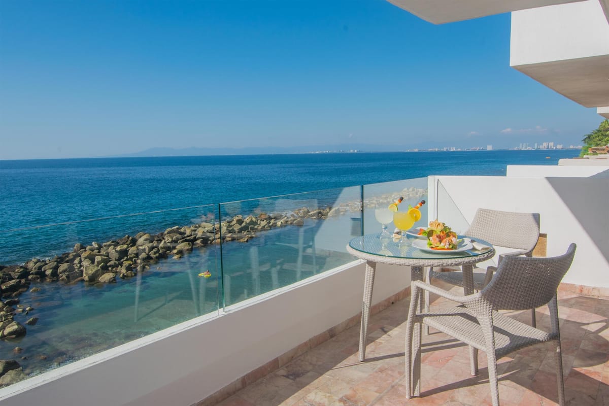Varanda com guarda-corpo de vidro com vista para a costa rochosa e o mar. Há uma mesa com cadeiras e tampo de vidro, e uma pequena planta sobre a mesa.