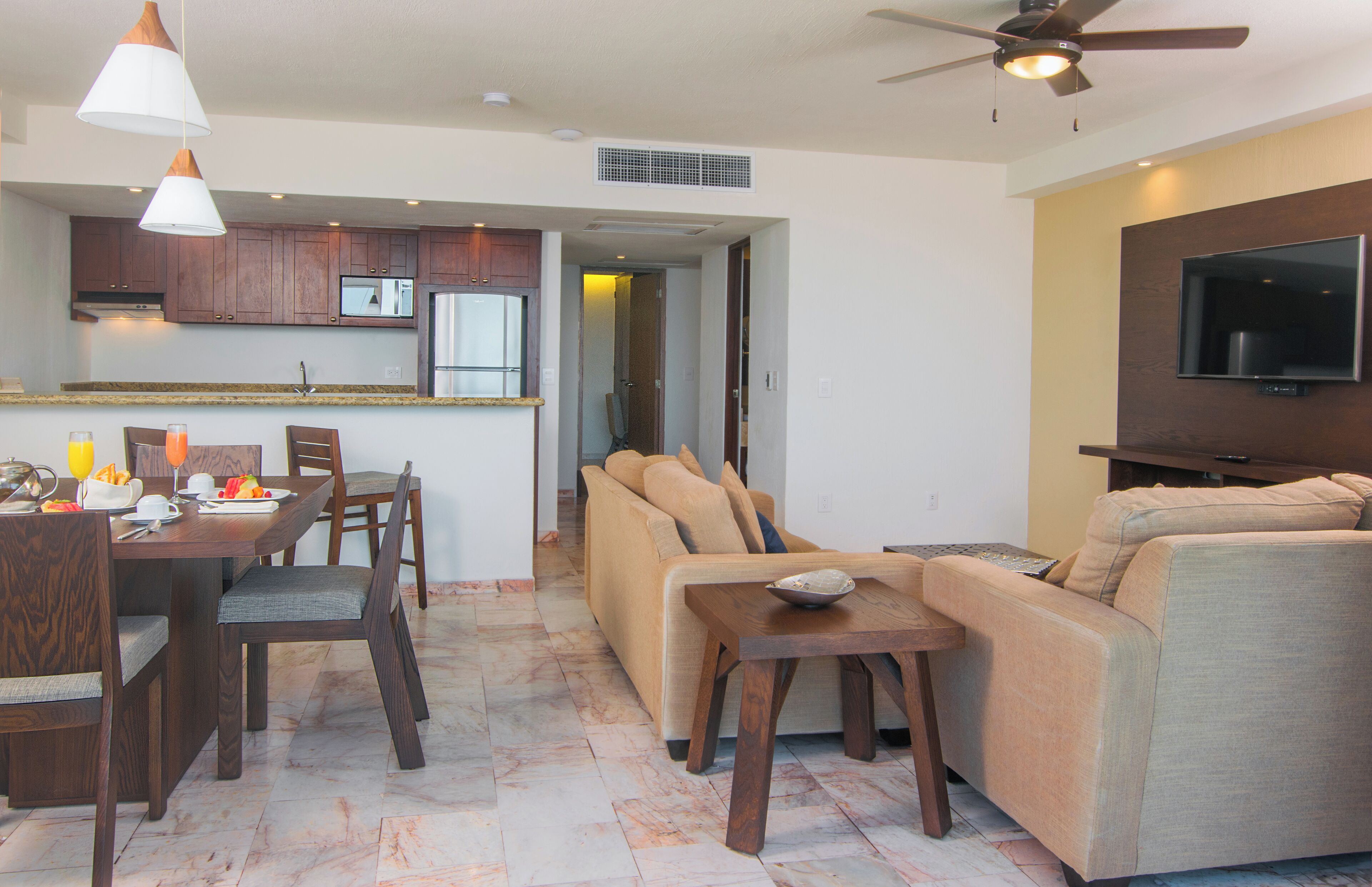 A modern living room with a sofa, dining table, and chairs. There is a kitchen area with cabinets and a microwave. A ceiling fan and a flat-screen TV are also visible.