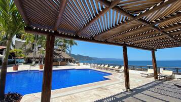 Zona de piscina con una pérgola de madera, palmeras y vista al mar.
