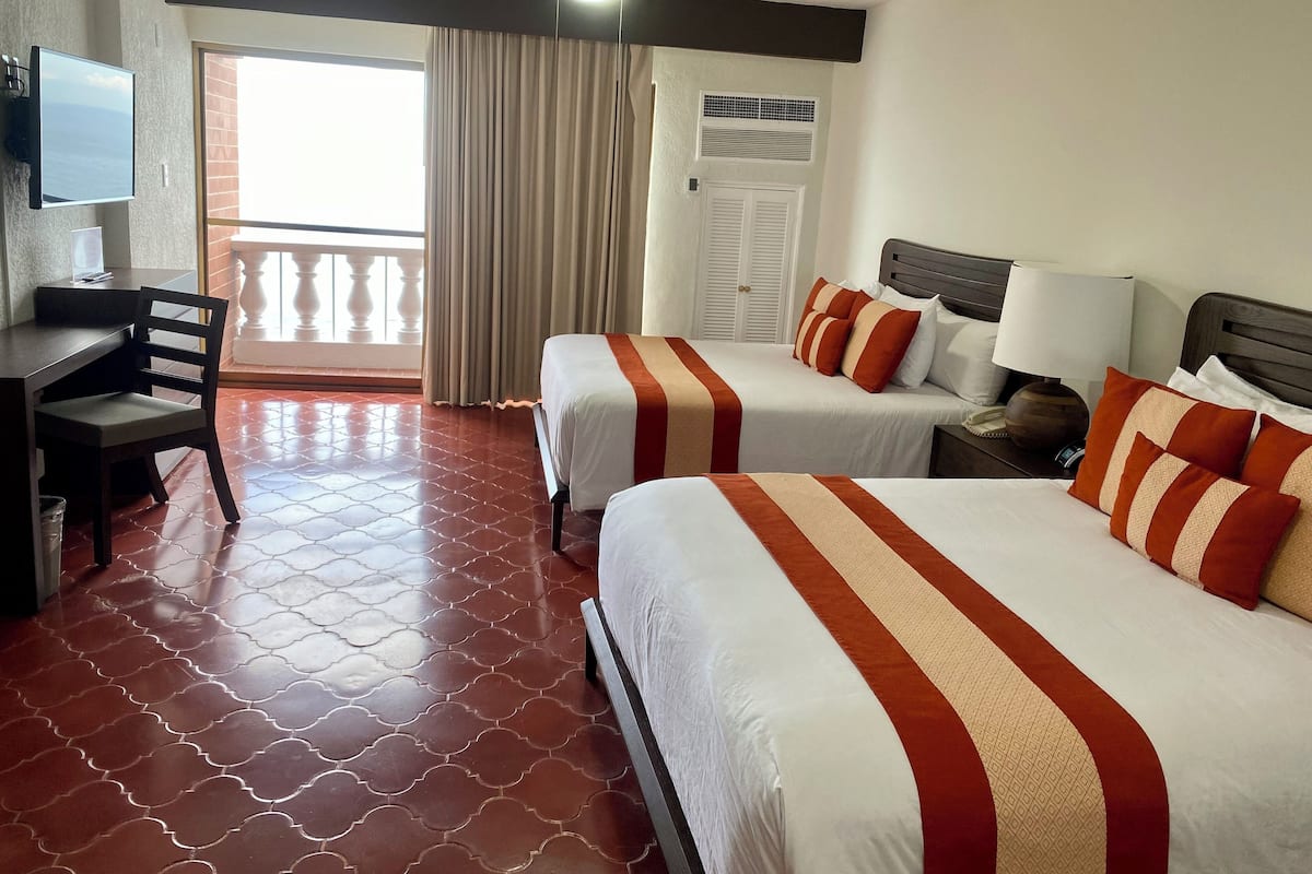 Quarto de hotel com duas camas, uma mesa, uma cadeira, uma TV e uma varanda com cortinas.