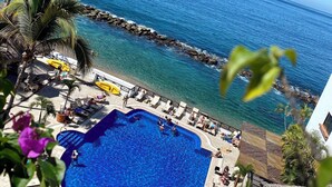 A pool area with clear blue water, surrounded by lounge chairs and umbrellas, with a view of the ocean and a rocky shoreline. - Costa Sur All Inclusive Classico Collection by Sonesta (Puerto Vallarta)