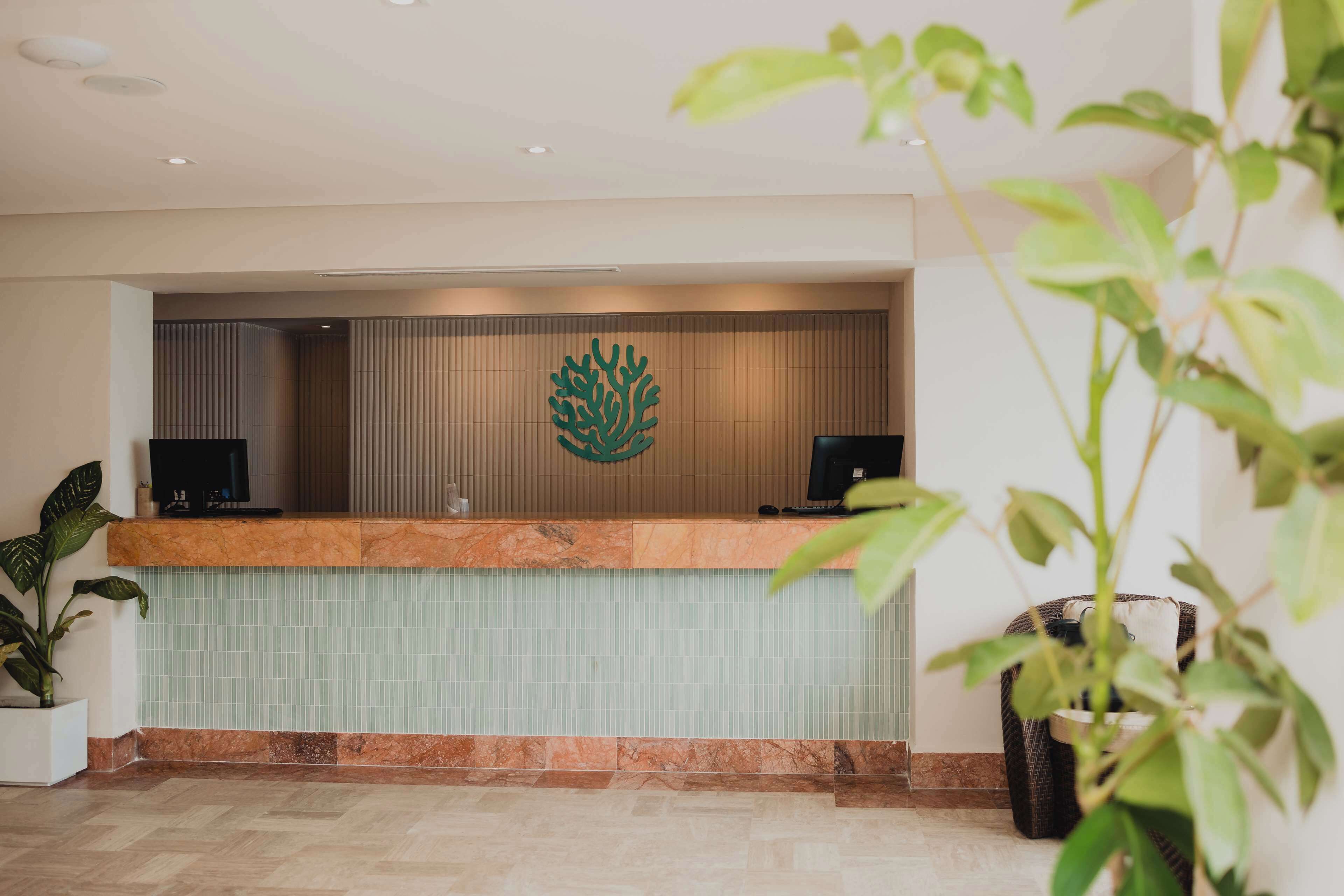 A reception desk with a green logo, two monitors, and a tiled wall.