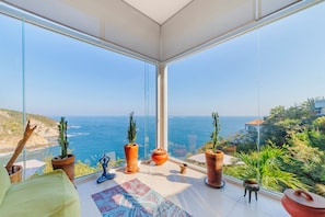A modern room with large glass windows overlooking a coastal view, featuring potted plants and a yoga mat.