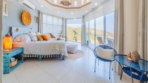 Un dormitorio amplio con una cama grande, una mesita y una silla. Cuenta con un balcón con vistas a las colinas.
