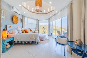 Une chambre spacieuse avec un grand lit, une petite table et une chaise. Elle dispose d’un balcon avec vue sur les collines.