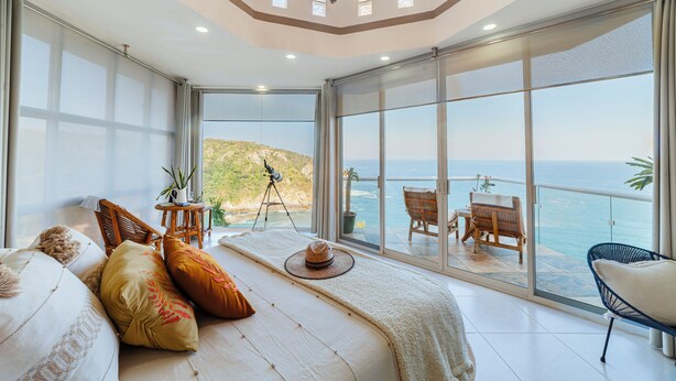 Un dormitorio con un ventanal grande con vistas al mar, un balcón con mobiliario exterior y una lámpara de techo.