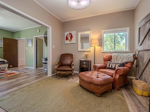 A living room with a brown leather sofa, a matching ottoman, a wooden side table, a floor lamp, and a window with a view of trees. - Fruita Finds: Dog Friendly Downtown Gem (Fruita)