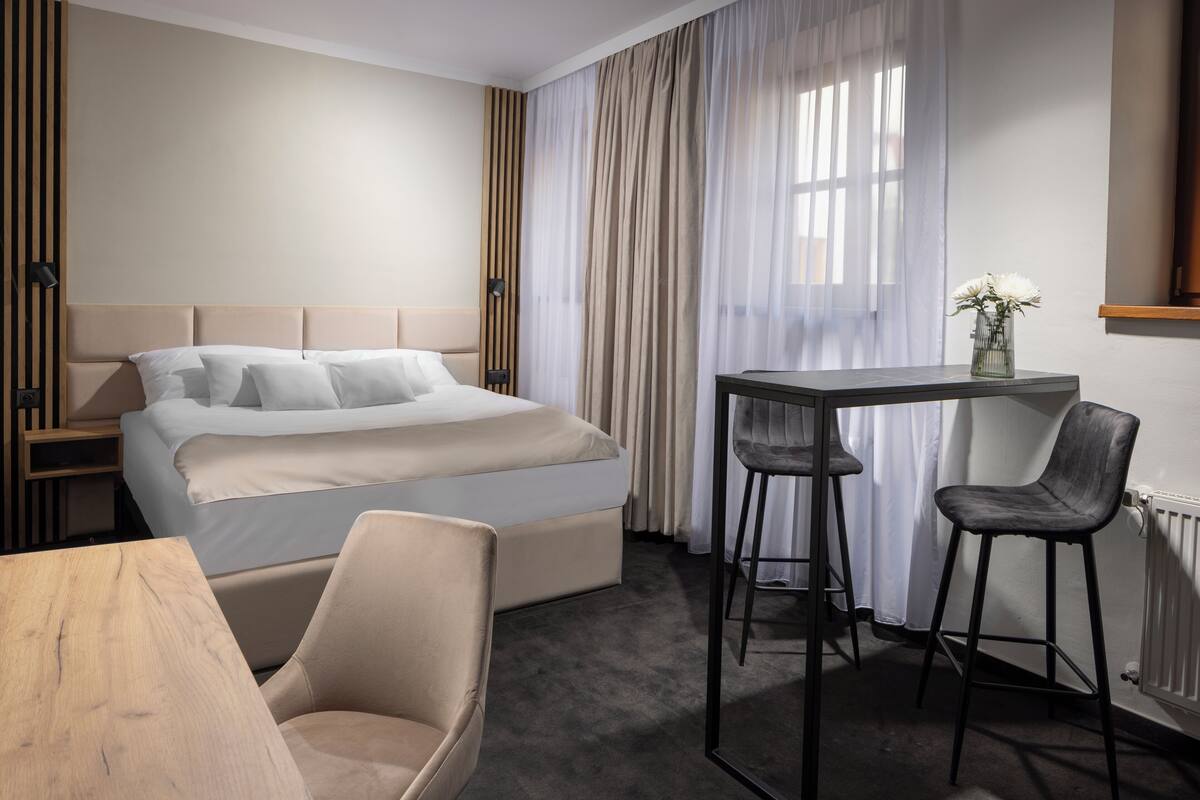 A modern hotel room with a large bed, a small table with two chairs, a wooden desk, and a mirror on the wall.