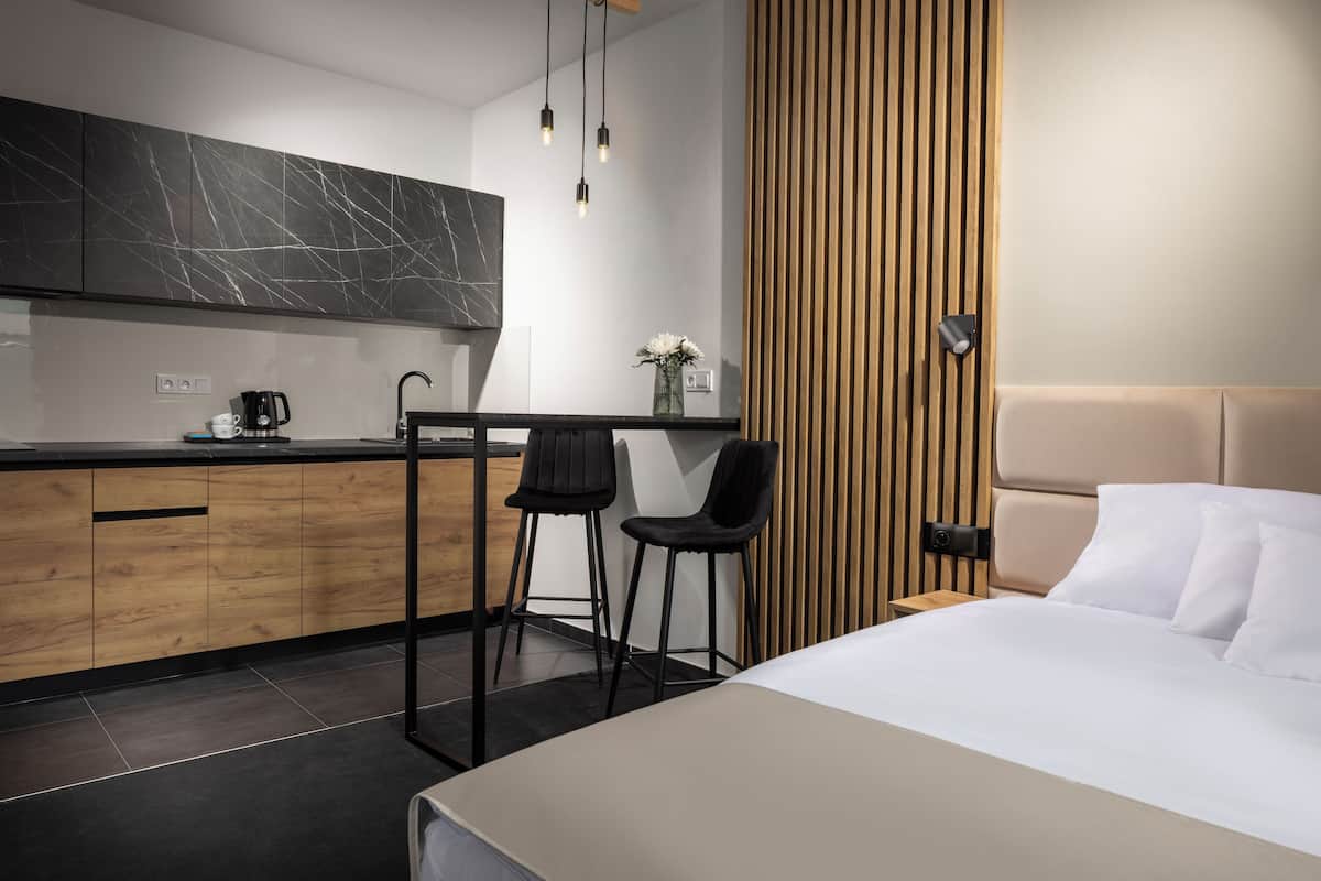 A modern hotel room with a bed, a small kitchen area with a marble backsplash, and a wooden panel wall.