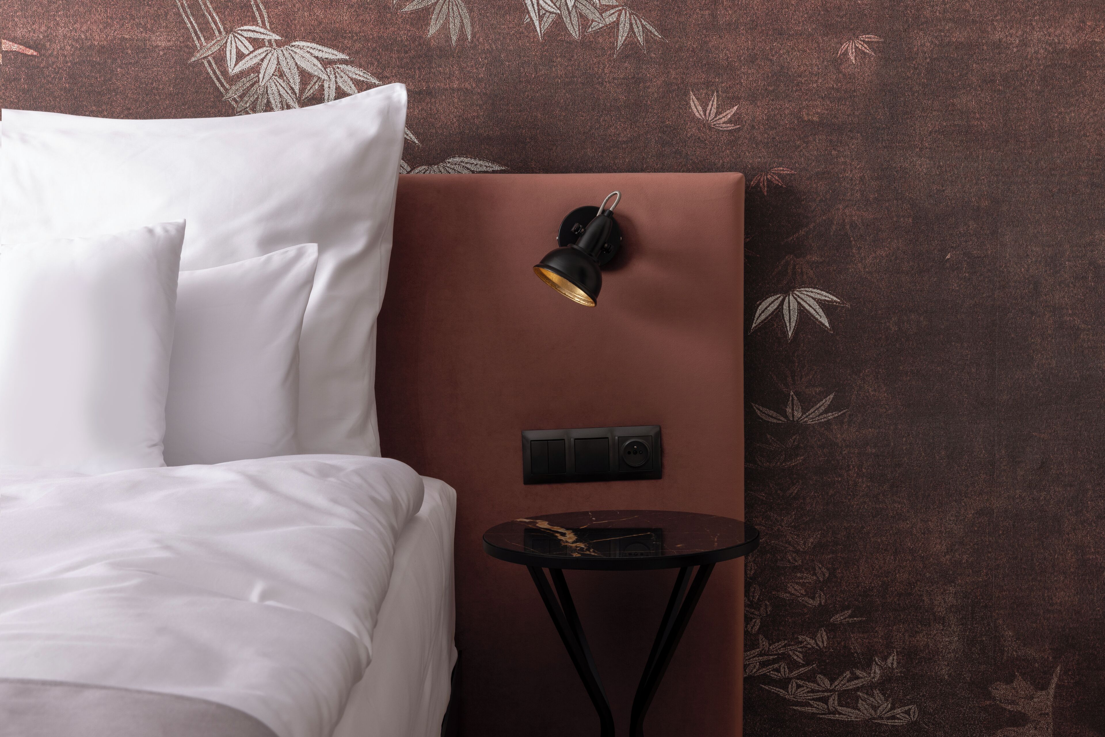Een opgemaakt bed met witte lakens, een klein rond tafeltje met een zwarte blad en een wandlamp.