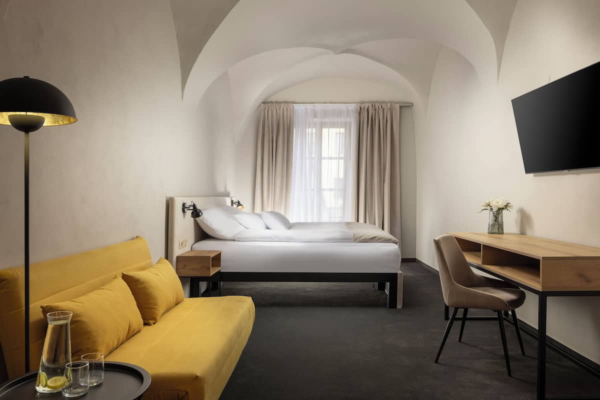 A hotel room with a bed, a desk, a chair, a sofa, a lamp, and a TV on the wall.