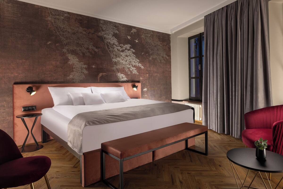 A modern bedroom with a large bed, a bench, a red chair, a small table, and a wall with a leaf pattern.