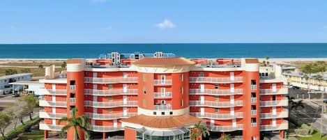 A multi-story hotel with a central courtyard, surrounded by a parking area and a beach view.