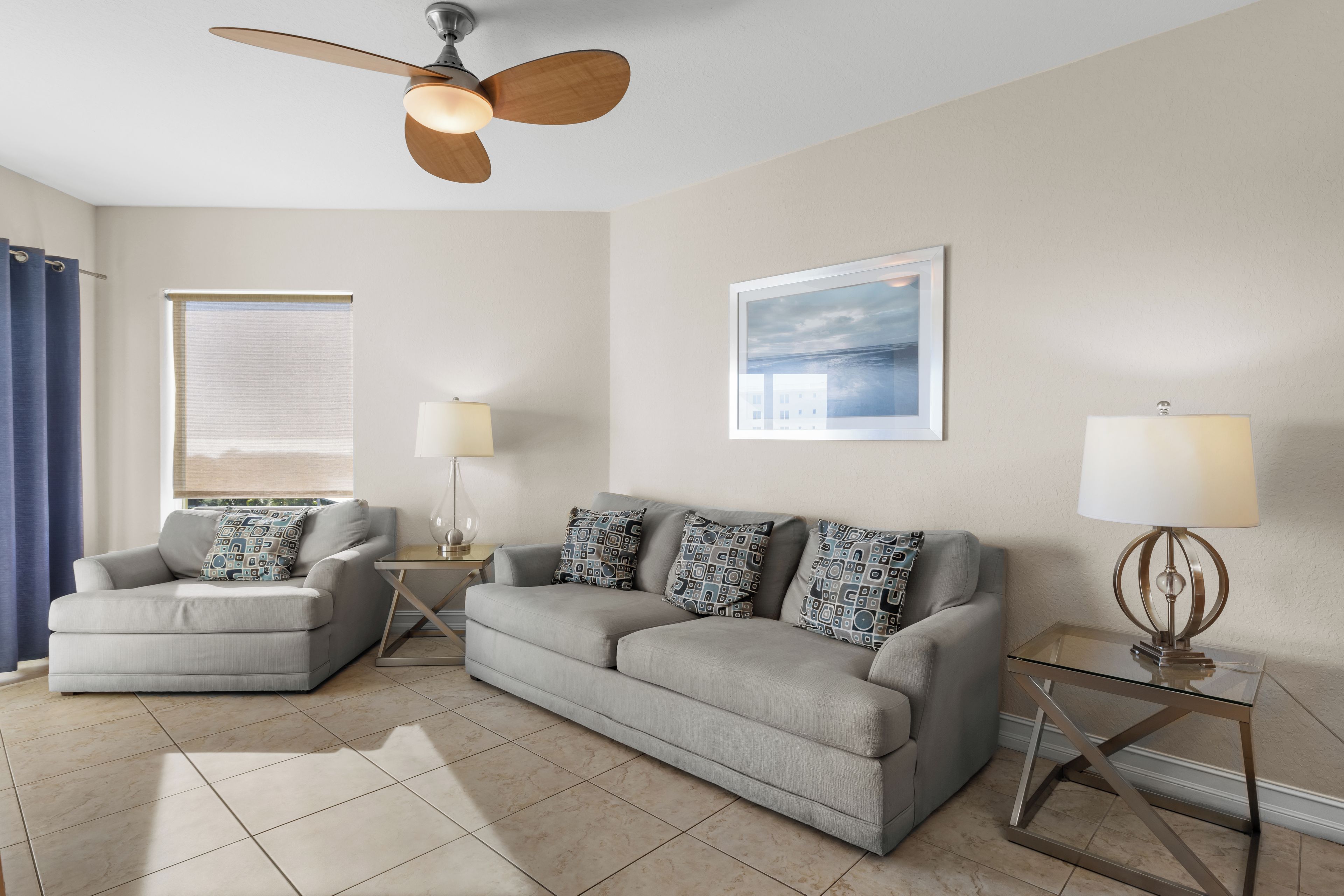 A living room with a grey sofa, a grey armchair, a ceiling fan, a framed picture on the wall, and a window with a view.