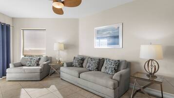 A living room with a grey sofa, a grey armchair, a ceiling fan, a framed picture on the wall, and a window with a view.