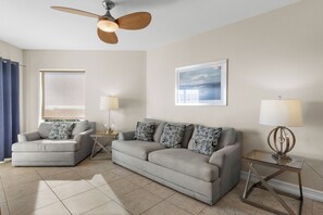 Un salon avec un canapé gris, un fauteuil gris, un ventilateur de plafond, un tableau encadré au mur et une fenêtre donnant sur un paysage.