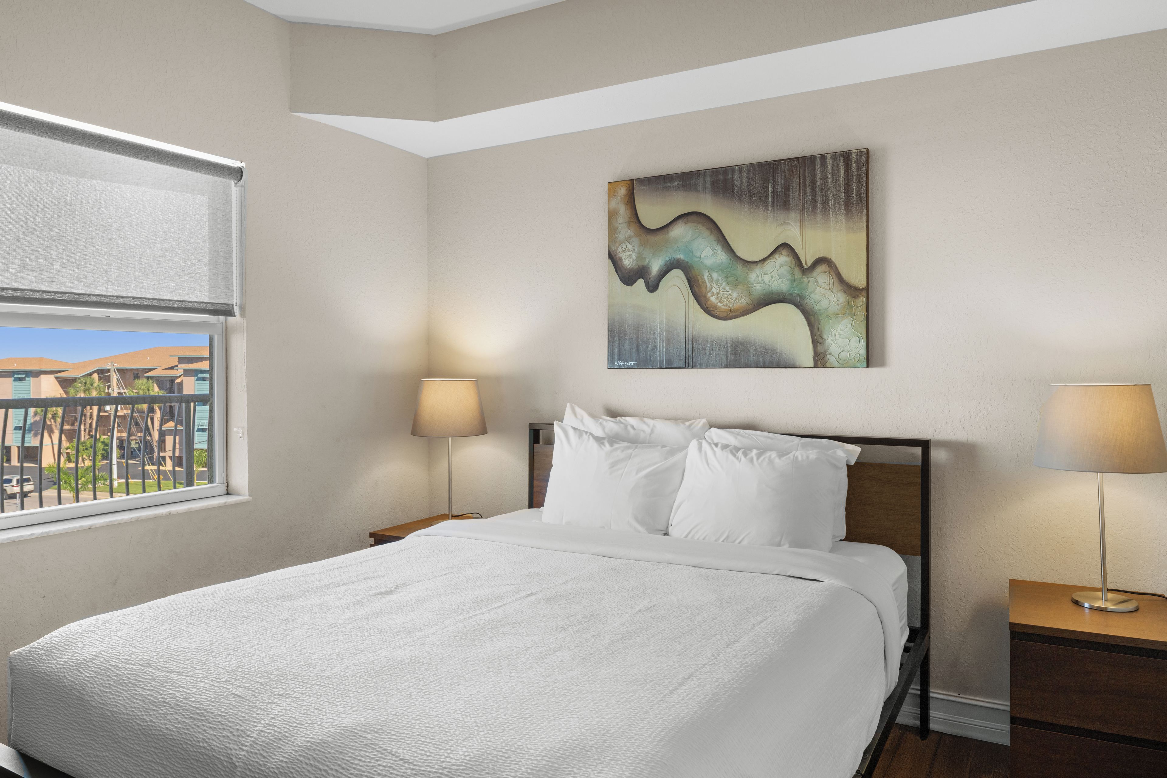 A hotel room with a bed, two bedside lamps, a painting on the wall, and a window with a view of buildings.